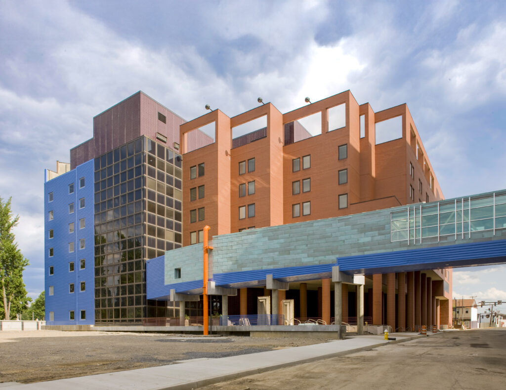 A modern building complex with red brick, blue metal siding, and a glass-enclosed skybridge connecting two structures.