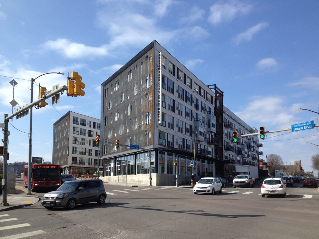 A city intersection with modern apartment buildings, a red bus, and several cars under a blue sky.