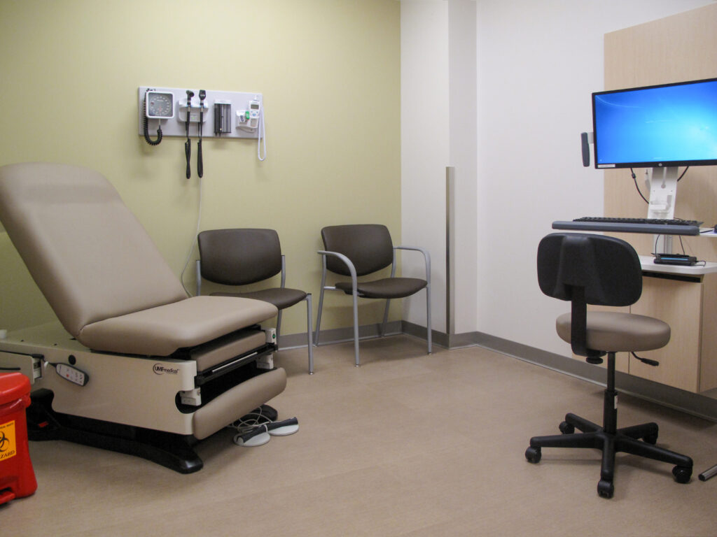A medical examination room with an adjustable exam table, two chairs, a wall-mounted diagnostic station, and a computer workstation.