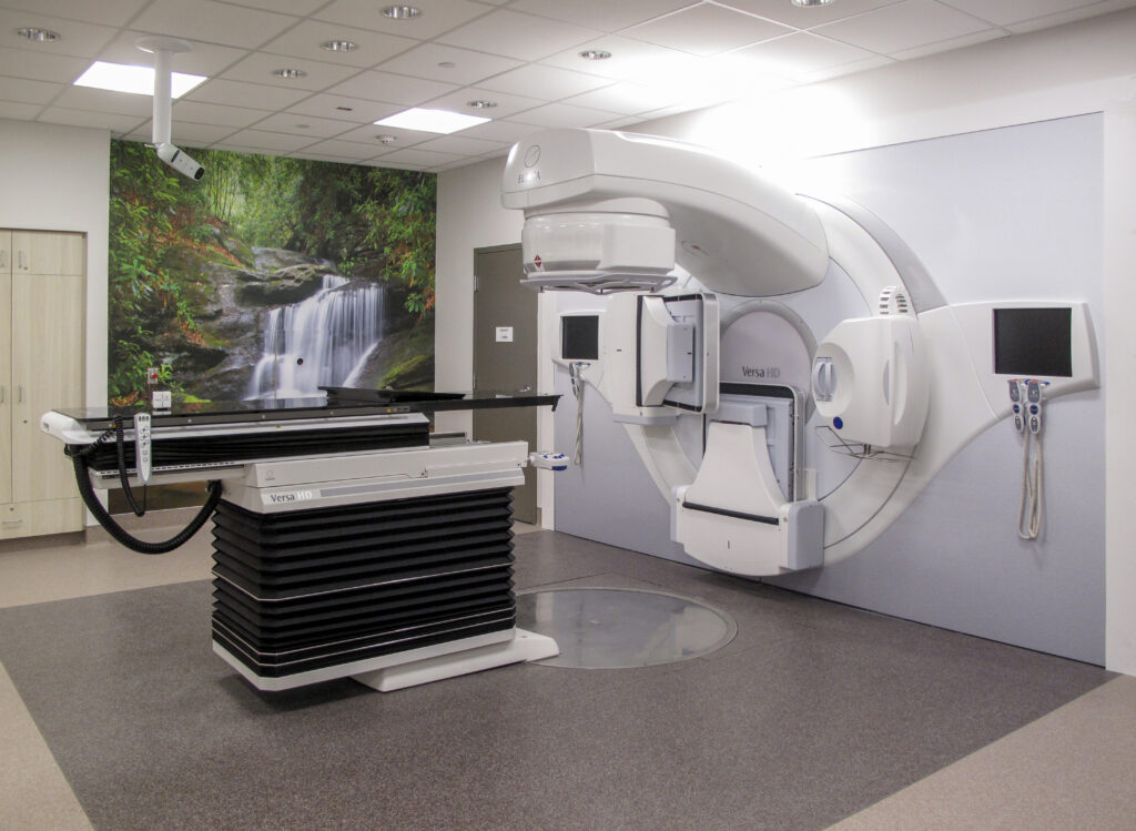 A medical linear accelerator for radiation therapy in a treatment room with a wall mural of a waterfall.
