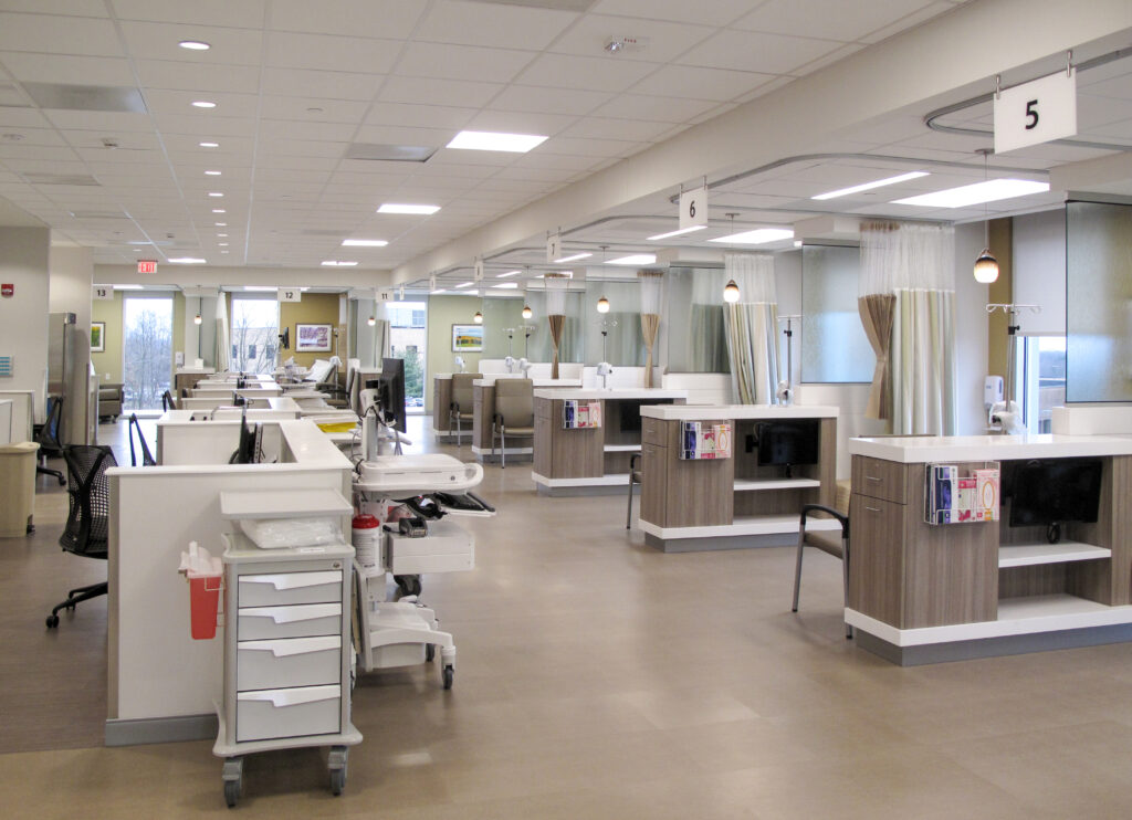 A modern medical infusion center featuring rows of partitioned patient bays, each with a reclined chair, monitor, and privacy curtain.