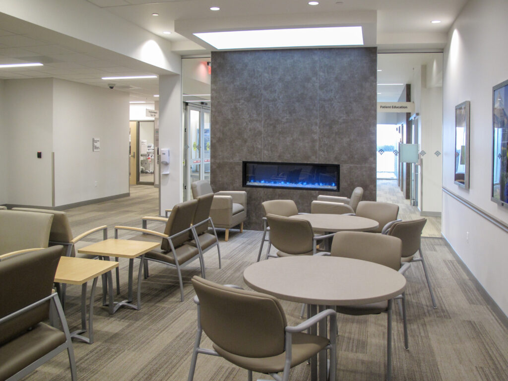 A hospital waiting room with beige chairs, small tables, and a linear fireplace built into a dark gray feature wall.
