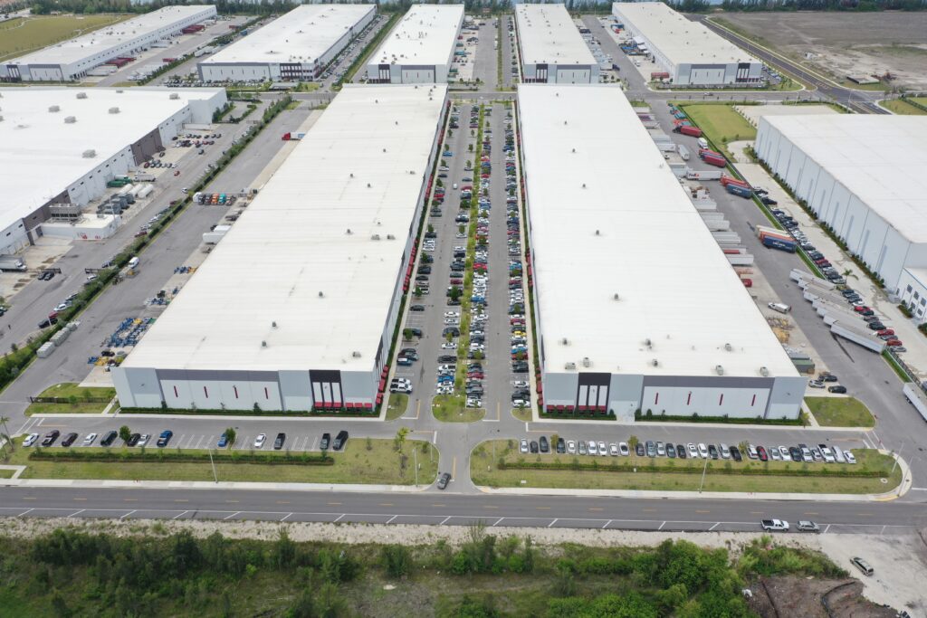 Aerial view of large industrial warehouse buildings with expansive parking lots filled with cars and shipping trucks.