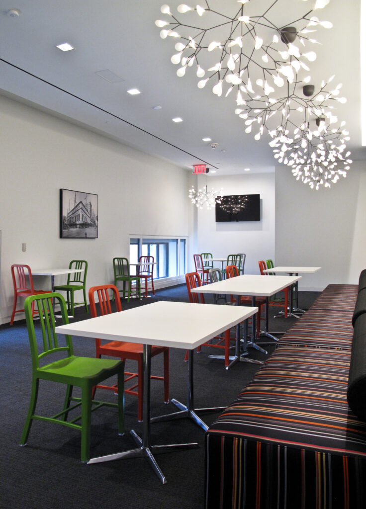 Modern office breakroom with white tables, colorful metal chairs, and long striped bench seating under contemporary branching chandeliers.