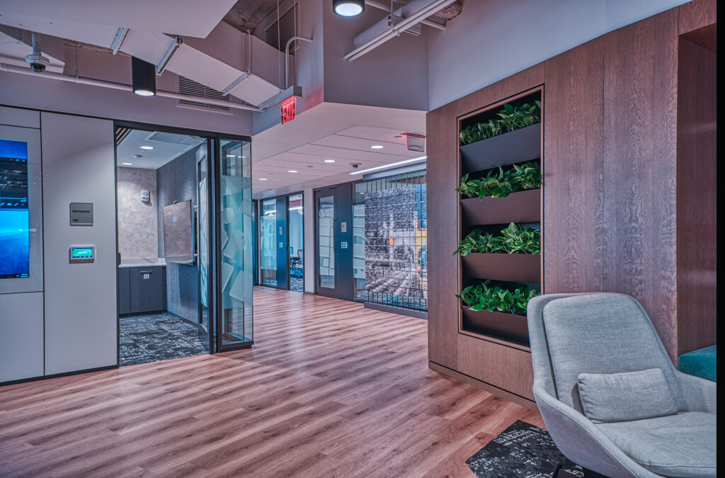 Modern office interior with wood-look flooring, glass-walled conference rooms, and a vertical wall planter filled with green plants next to a gray armchair.