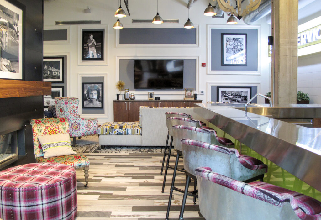 A lounge area featuring a stainless steel bar with stools, a flat-screen TV mounted above a sideboard, patterned chairs, and black-and-white framed photographs on the walls.