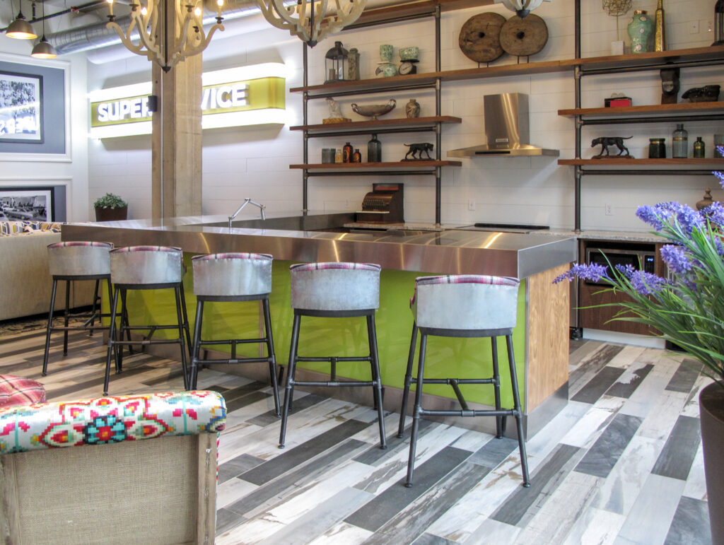 A stainless steel kitchen island with lime green panels and five bar stools, located in a room with industrial shelving and a “SUPER SERVICE” sign.