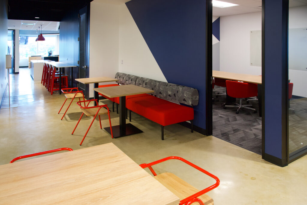 Office break area featuring red benches with patterned back cushions, light wood tables, and red metal chairs, with a glass-walled conference room in the background.