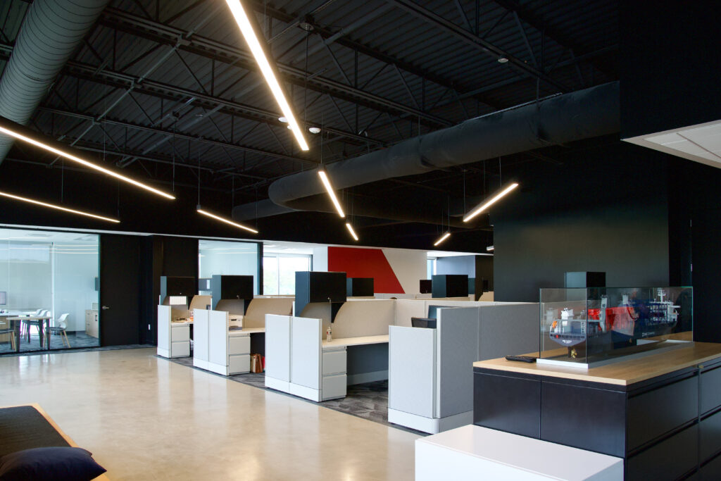 Modern open-plan office with grey cubicle workstations, a dark exposed ceiling, and a model ship in a glass display case.