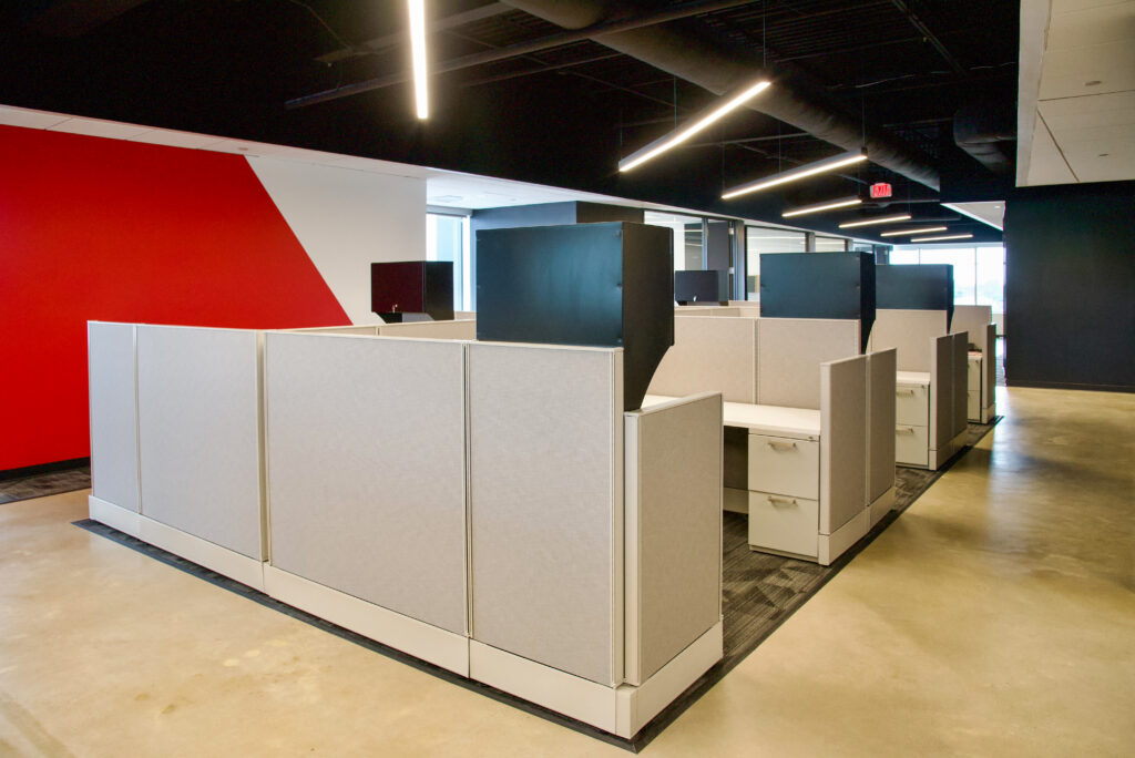 Rows of office cubicles with light gray partitions in a modern office space with concrete floors and a red accent wall.