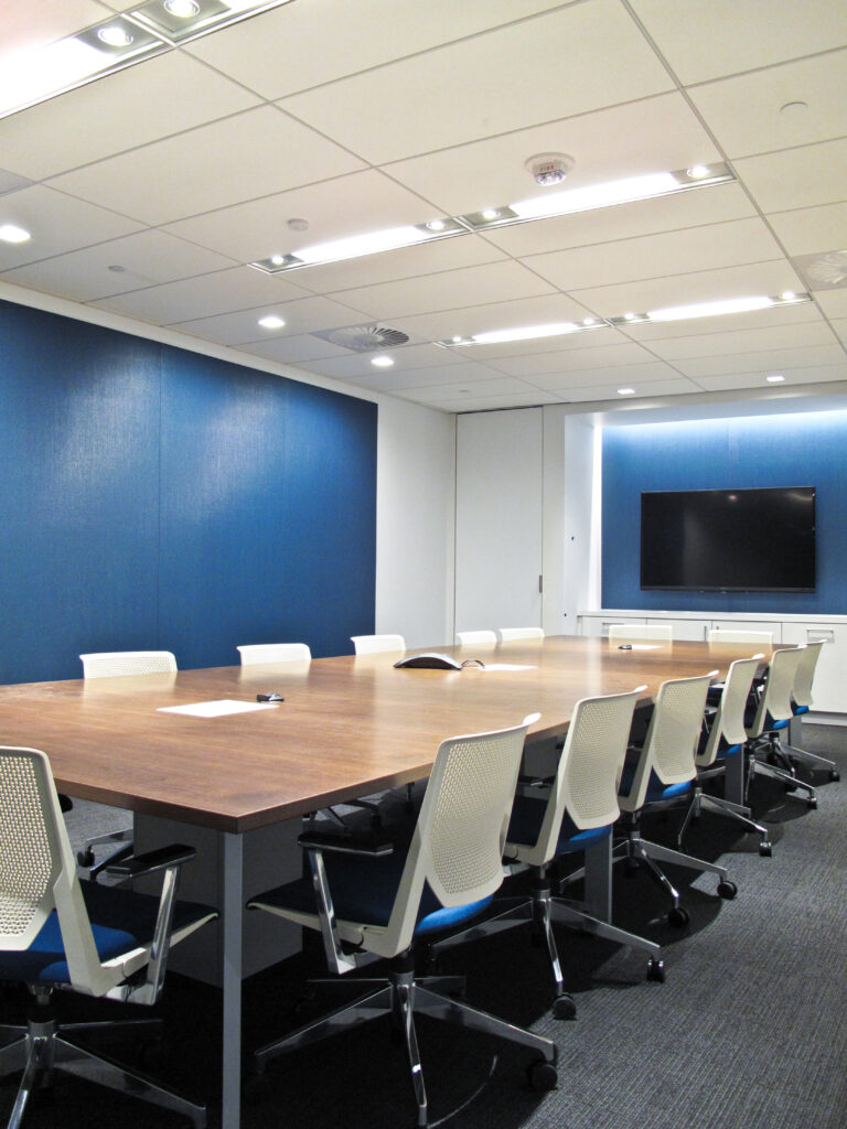 A modern conference room with a long wooden table, white office chairs, and a wall-mounted flat-screen television.