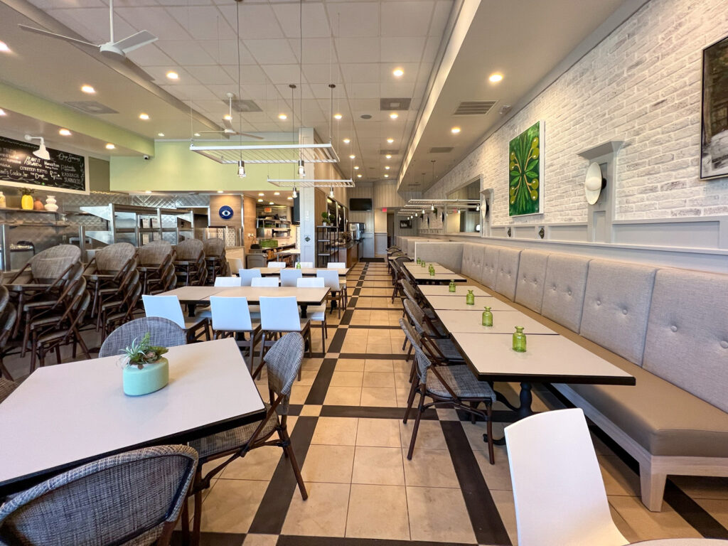 Empty restaurant dining room with a mix of tables, chairs, and bench seating, featuring a white brick wall and tile floor.