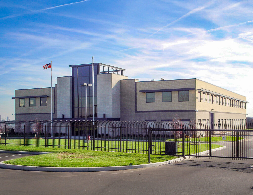 A two-story commercial building with light brick walls and a central glass facade, enclosed by a black metal fence.