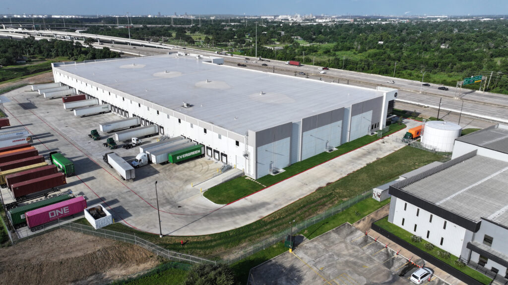 Aerial view of a warehouse with multiple loading docks, shipping containers, and trucks parked alongside an adjacent highway.
