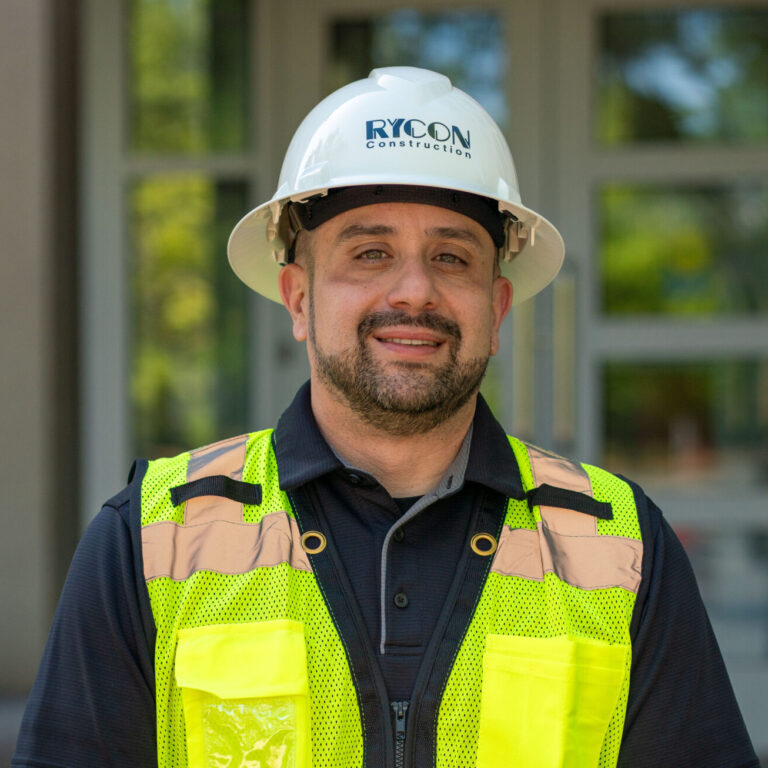 A construction worker wearing a white Rycon Construction hard hat and a high-visibility yellow safety vest.