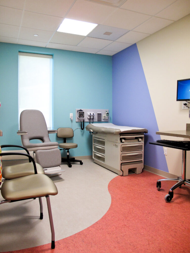 A doctor’s examination room with a medical exam table, a patient chair, a stool, and a computer workstation.
