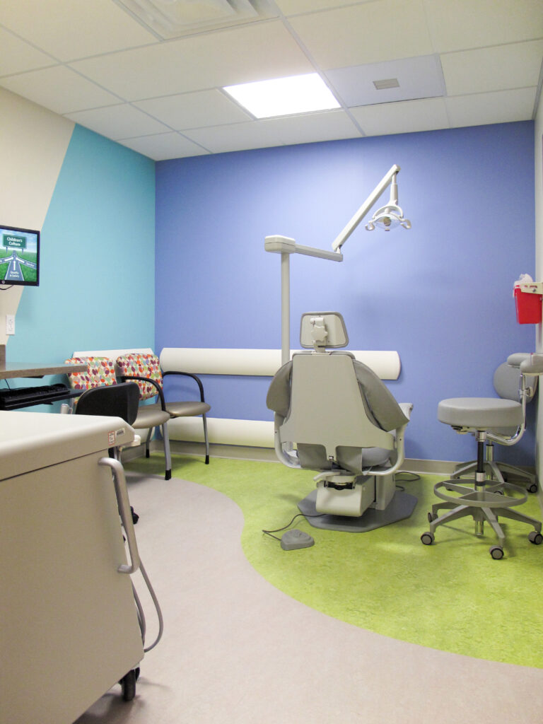 An empty dental exam room with a patient chair, a rolling stool, and two colorful chairs against blue and teal walls.