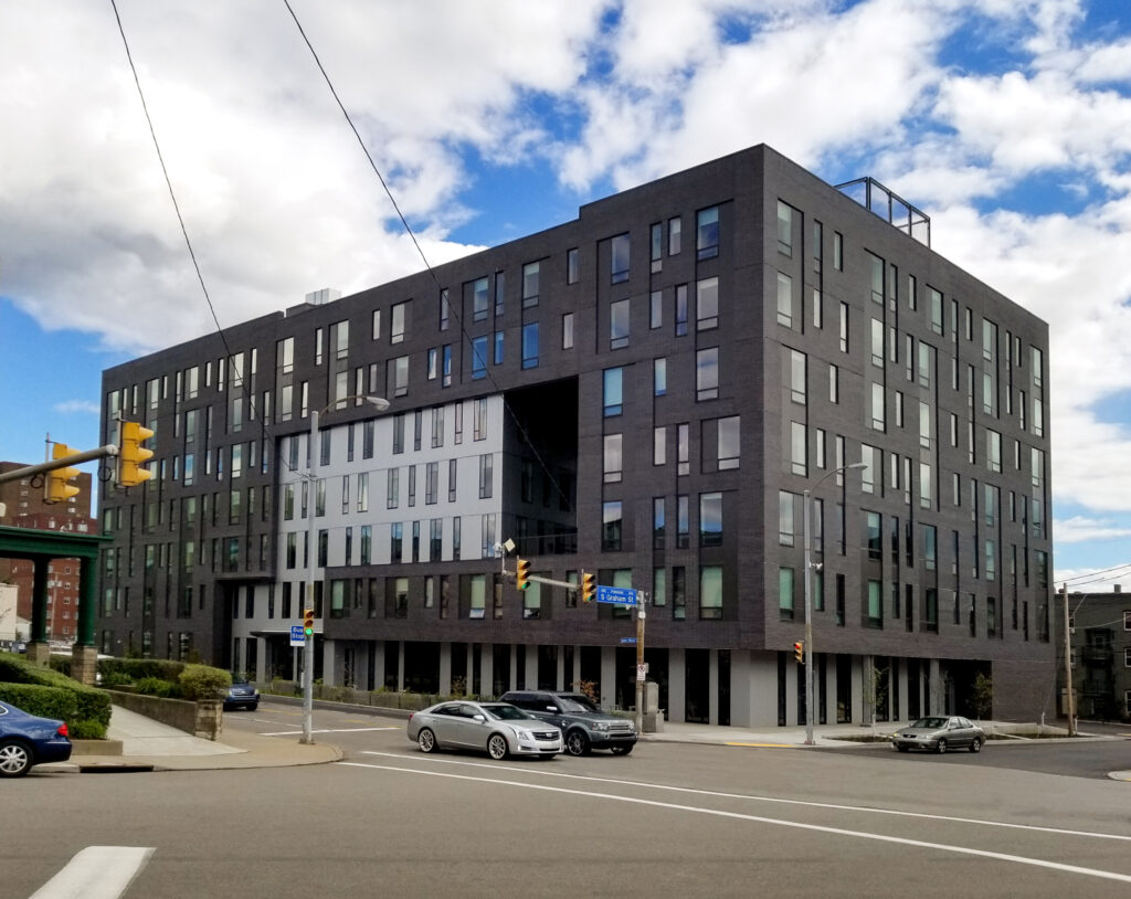 A large, modern building with a dark brick exterior and a central recessed section, located on a city street corner with traffic.