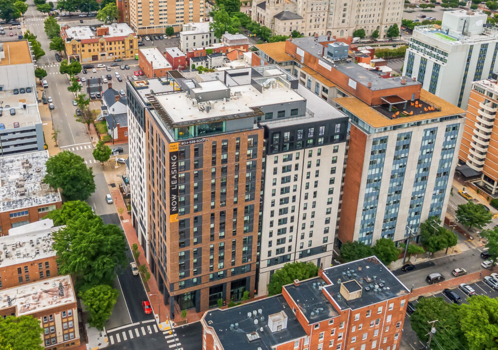 Aerial view of a mid-rise apartment building with a “Now Leasing” sign and phone number on the side, surrounded by city streets and other buildings.