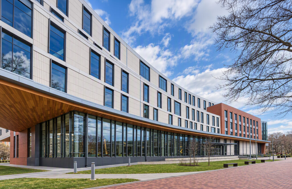 Modern multi-story building with a curved glass and tan panel facade, featuring a wooden ceiling overhang and a brick walkway.