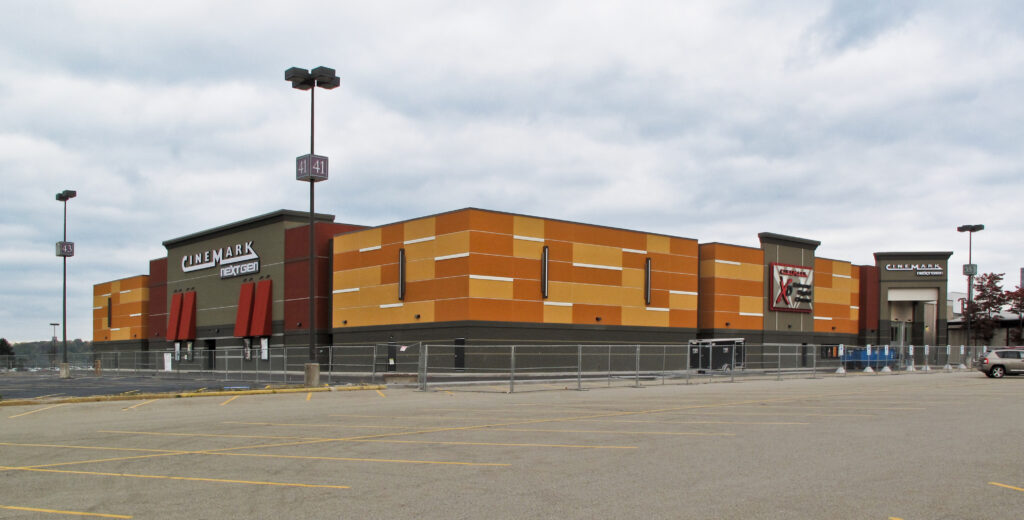 A Cinemark theater building featuring a yellow, white, and dark gray facade with “NextGen” and “XD” signage, situated in a large empty parking lot.