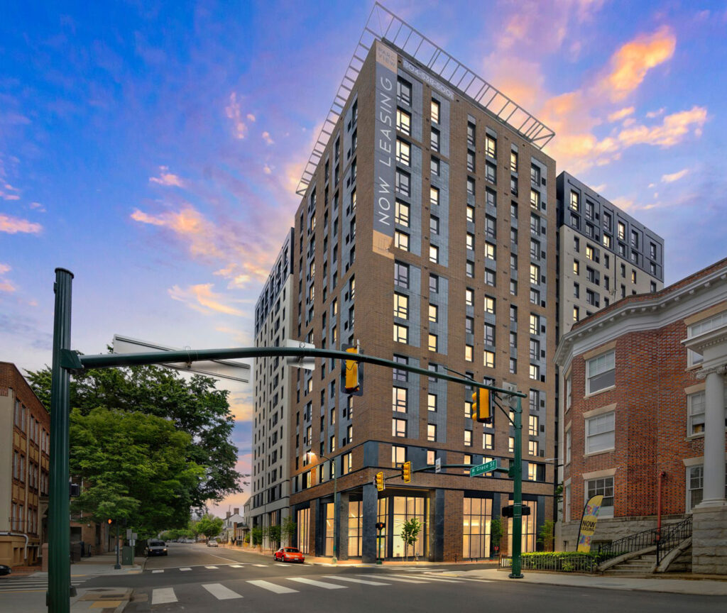 A modern high-rise apartment building at a city intersection at dusk, with a “Now Leasing” banner on the side.