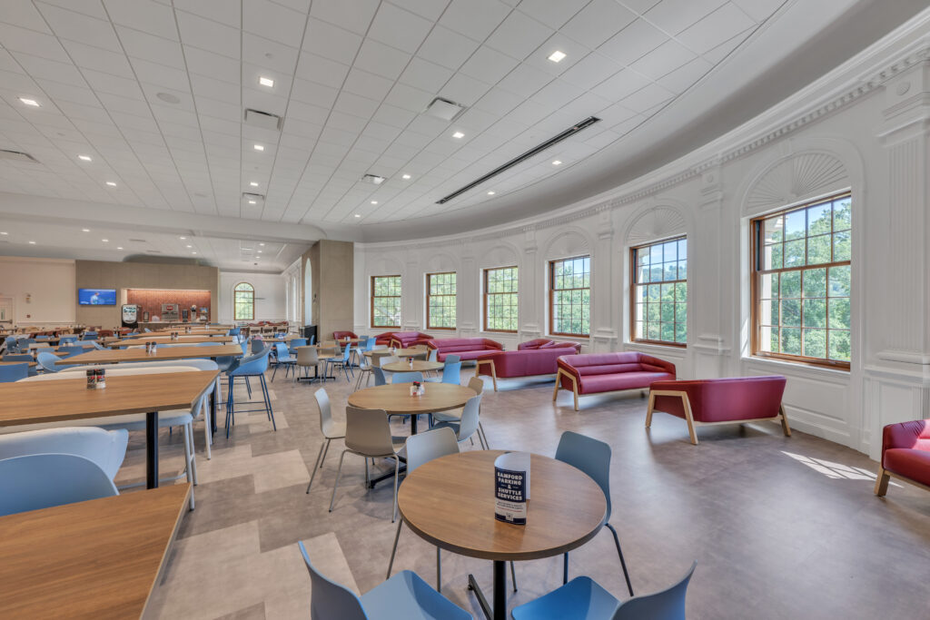 A large, sunlit lounge with various round and long tables, blue chairs, and red sofas arranged along a wall of arched windows.