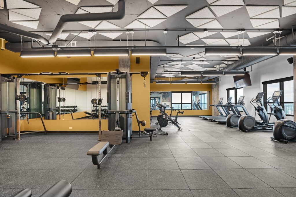 A modern fitness center featuring a cable machine, exercise bikes, and treadmills in front of a yellow wall with mirrors.