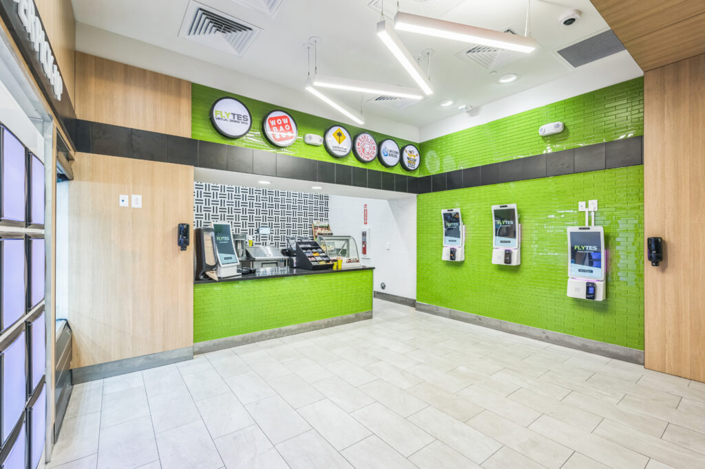 A fast-casual dining hall with a service counter, Flytes signage, and three self-service ordering kiosks mounted on a bright green tiled wall.