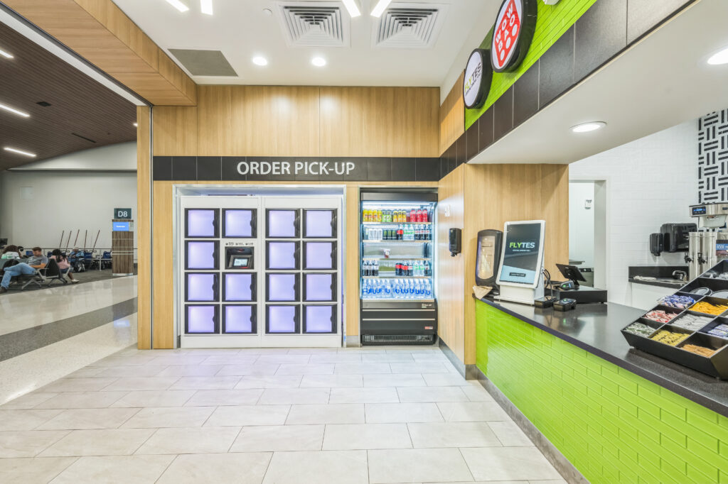 Airport cafe counter featuring an order pickup kiosk with lockers, a refrigerated drink display, and a self-service ordering station.