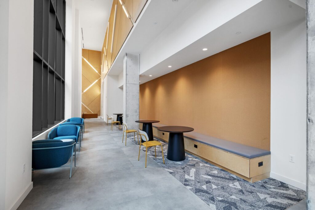 A long, narrow modern lounge area with rows of blue armchairs on the left, and a wooden bench with small tables and chairs on the right.