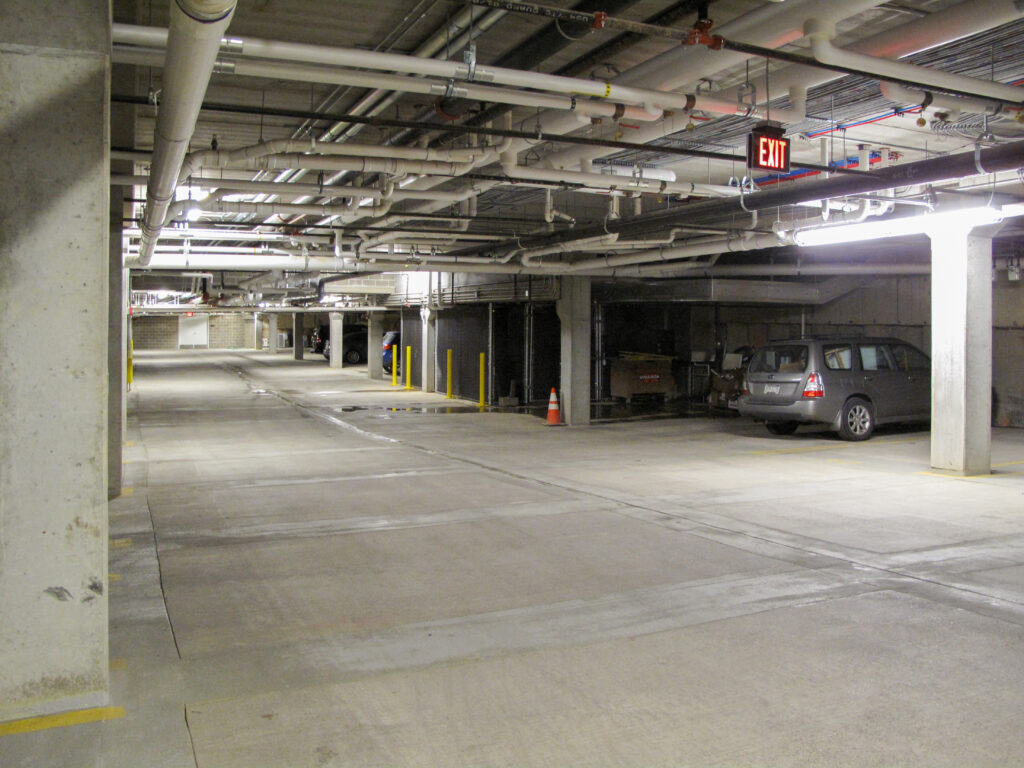 An indoor parking garage with concrete pillars, overhead pipes, an illuminated exit sign, and a silver minivan parked on the right.