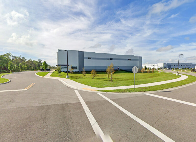A gray industrial warehouse building with the number 6135 on the corner, situated at an intersection with paved roads and grassy lawns.