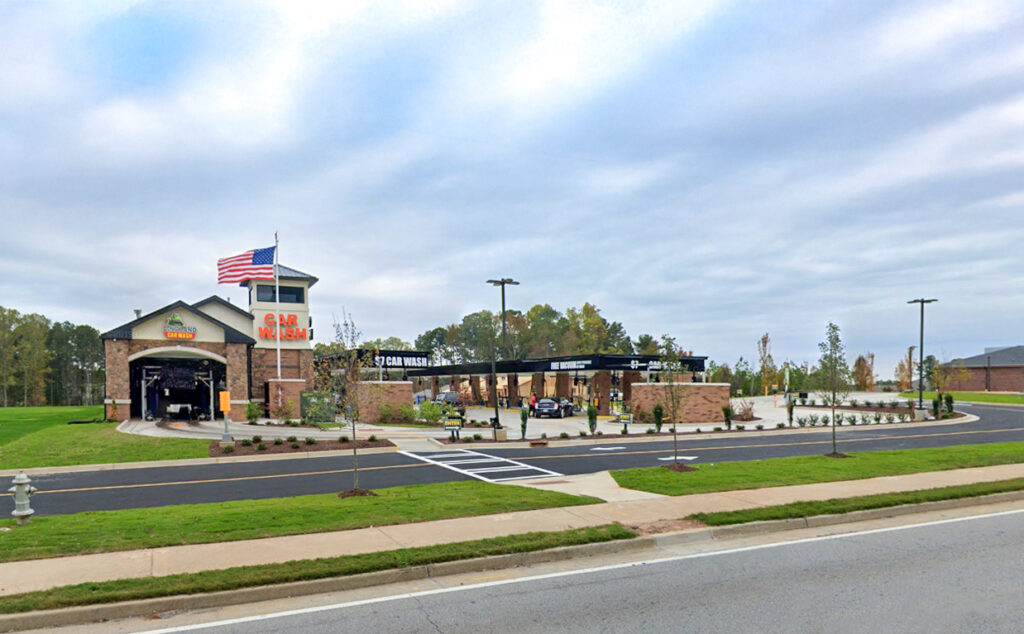 An exterior view of a drive-through car wash facility featuring a brick building with a large American flag and an adjacent vacuuming station.