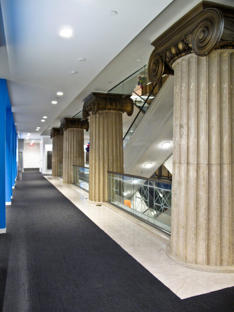 A hallway lined with tall, decorative fluted columns featuring ornate capitals next to an escalator and glass railing.