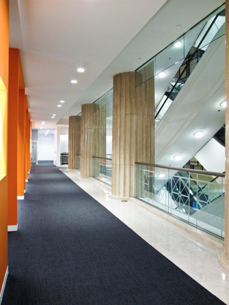 Long hallway with a dark carpet, orange pillars on the left, and a glass-paneled escalator on the right.
