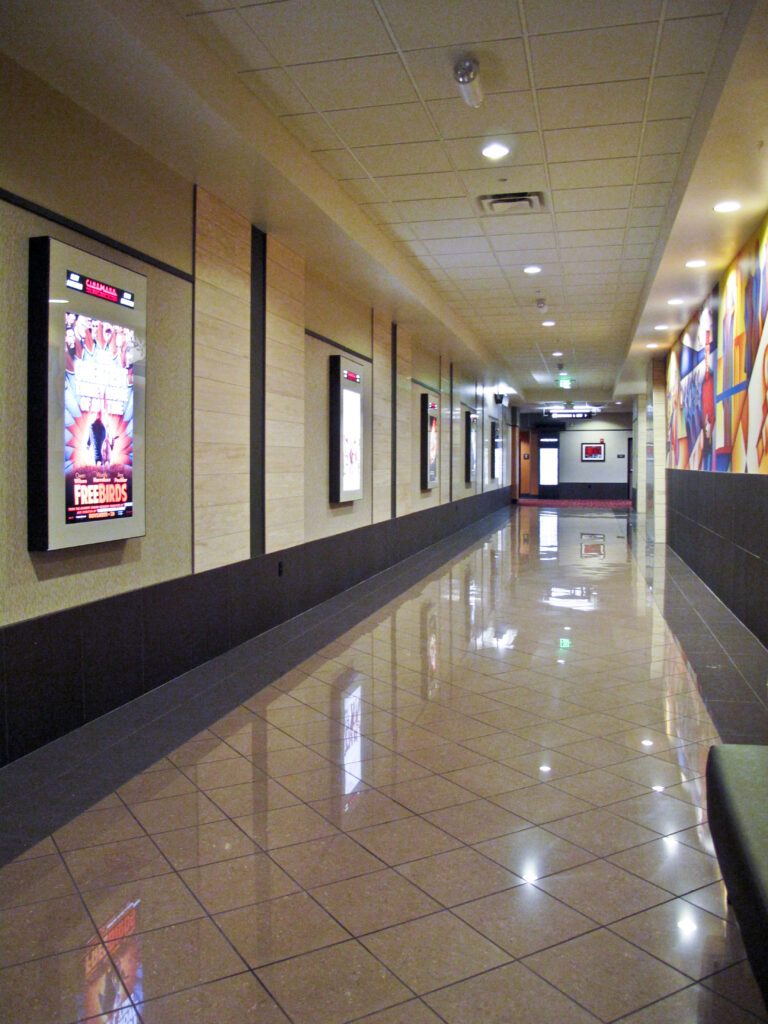 A long hallway in a movie theater with illuminated film posters on one wall, a mural on the other, and a polished tiled floor.