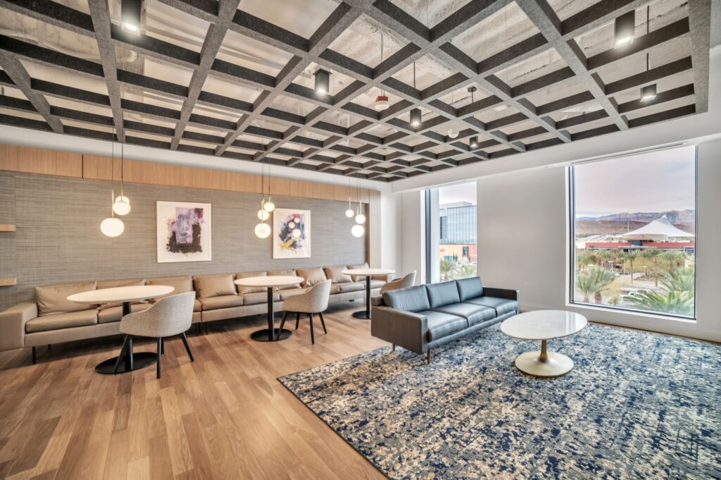 Lounge area with a grid ceiling, leather benches, small dining tables, a sofa, and a large window view of a parking lot and mountains.