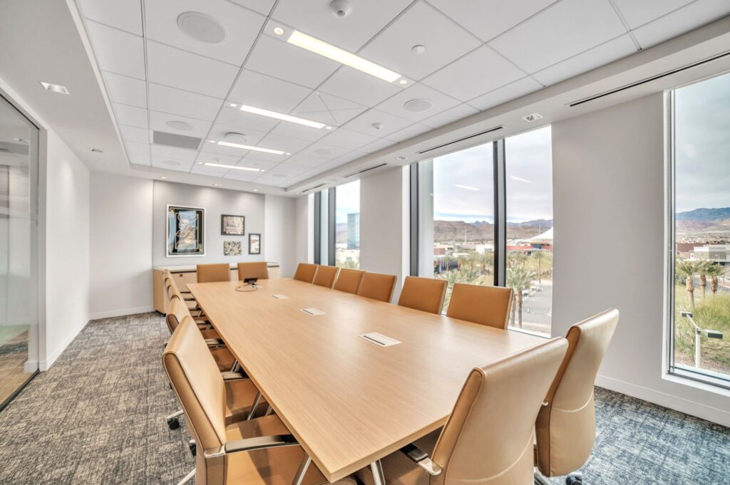 A long rectangular wooden conference table surrounded by ten tan office chairs in a modern meeting room with large windows overlooking a desert landscape.