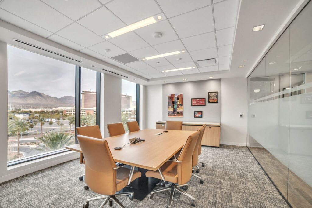 A conference room with a large wooden table, six tan office chairs, and framed artwork on the wall, overlooking a desert city skyline.