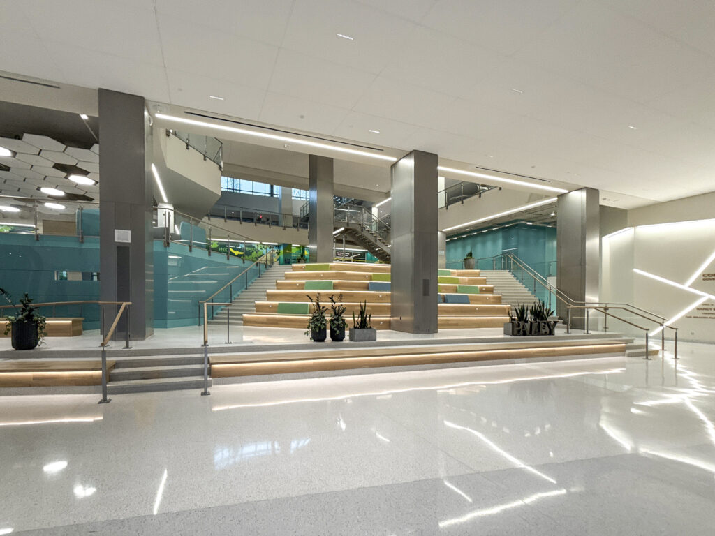 A wide, modern indoor atrium with tiered wooden seating steps, teal accents, and potted plants.