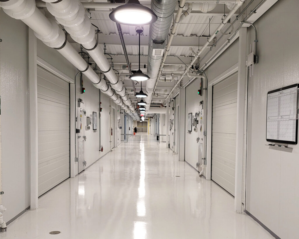 A long, bright industrial hallway with white walls, white roll-up doors, and exposed piping on the ceiling.