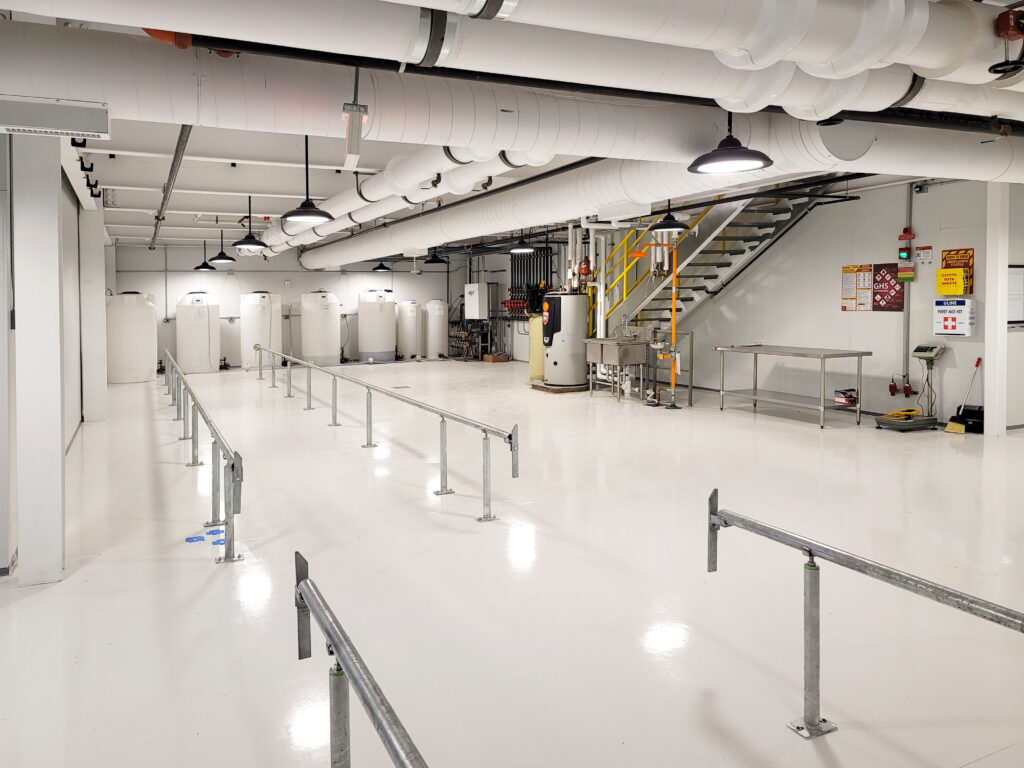 An industrial facility interior with white floors, several large white storage tanks, a staircase, and a metal work table.