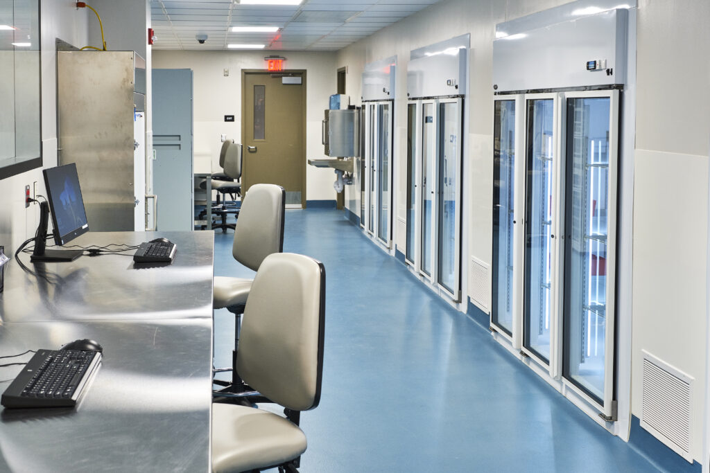 A laboratory hallway with stainless steel workstations and computers on the left, and a row of glass-fronted refrigeration units on the right.