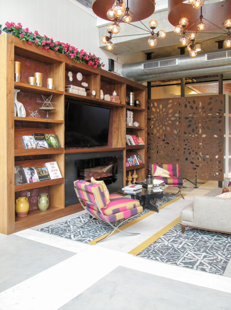 A modern lounge area with a built-in wooden shelving unit, a fireplace with a television, two striped armchairs, a gray sofa, and decorative laser-cut metal room dividers.