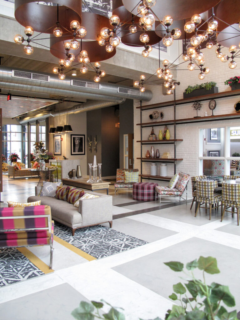 A modern hotel lobby with patterned armchairs, a grey sofa, and industrial-style tiered lighting fixtures hanging from the ceiling.