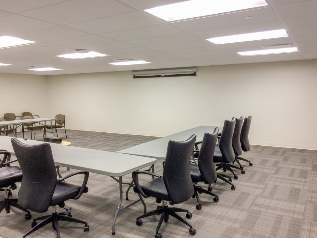 A conference room with modular folding tables arranged in a U-shape, surrounded by black office chairs on a carpeted floor.