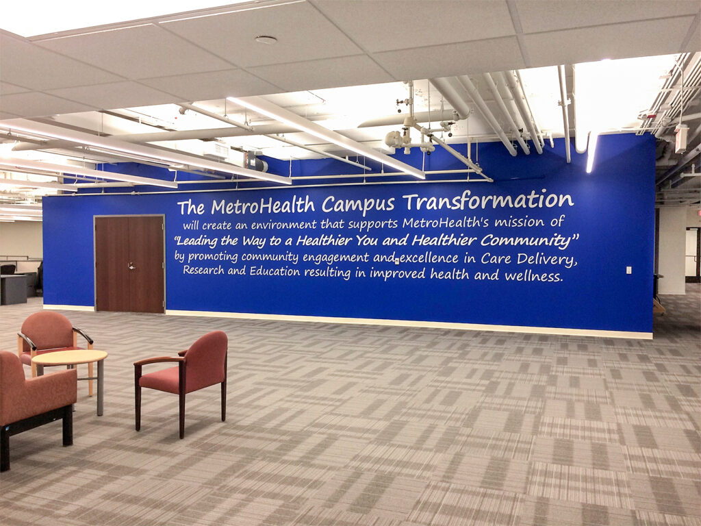 A blue wall in a lobby featuring white text describing The MetroHealth Campus Transformation, with two chairs and a side table in the foreground.