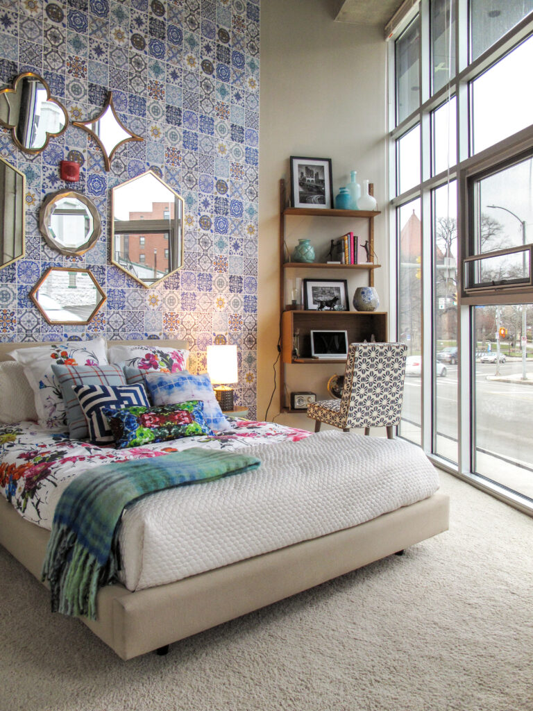 A bedroom with a patterned tiled accent wall, assorted wall mirrors, a bed with floral bedding, and a desk beside floor-to-ceiling windows.