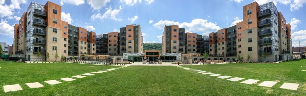 A wide panoramic view of a multi-story apartment complex surrounding a central courtyard with a large green lawn, outdoor seating areas, and a pool.